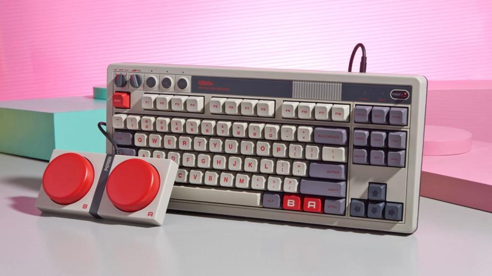 Die grau-rote mechanische Tastatur, genannt 8BitDo Retro Mechanical Keyboard, sitzt auf einem leuchtend rosa und pastellgrünen Hintergrund. Vor der Tastatur befindet sich ein riesiges rotes Paar